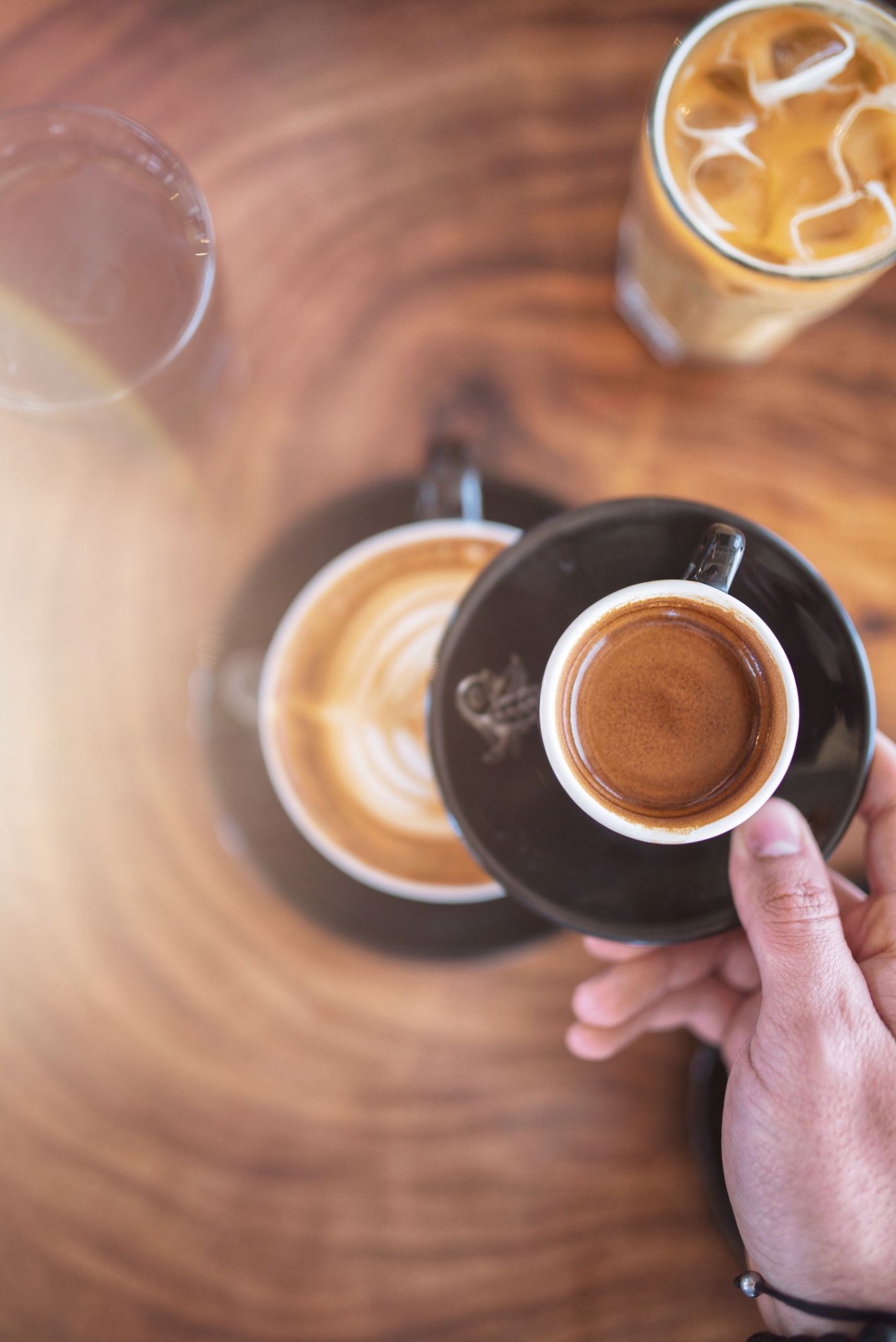 Ondergrond van hout daarop een glas met water, een glas ijskoffie een cappuccino en een hand die een espresso vasthoud.