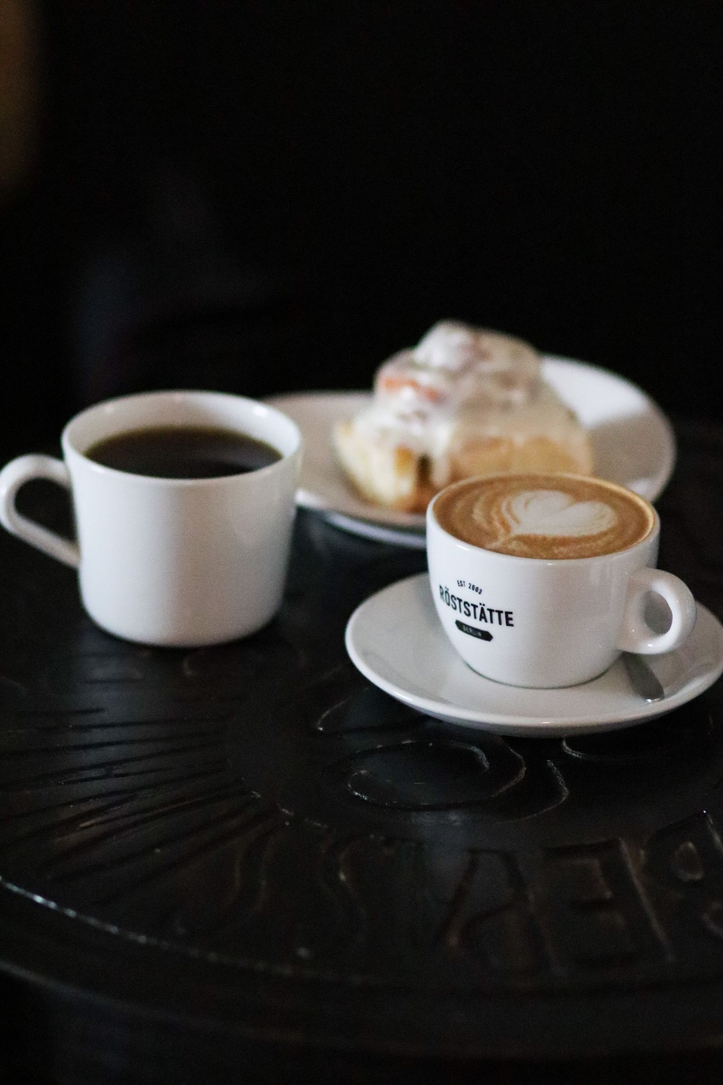 kopje zwarte koffie en een klein kopje cappuccino met daarachter een koffiekoek met geglazuurde suiker op een zwart tafeltje