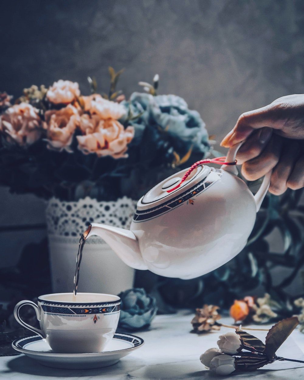 hand die thee aan het inschenken is met witte porseleinen thee pot in kopje. Romantische achtergrond met rozen in bloempot